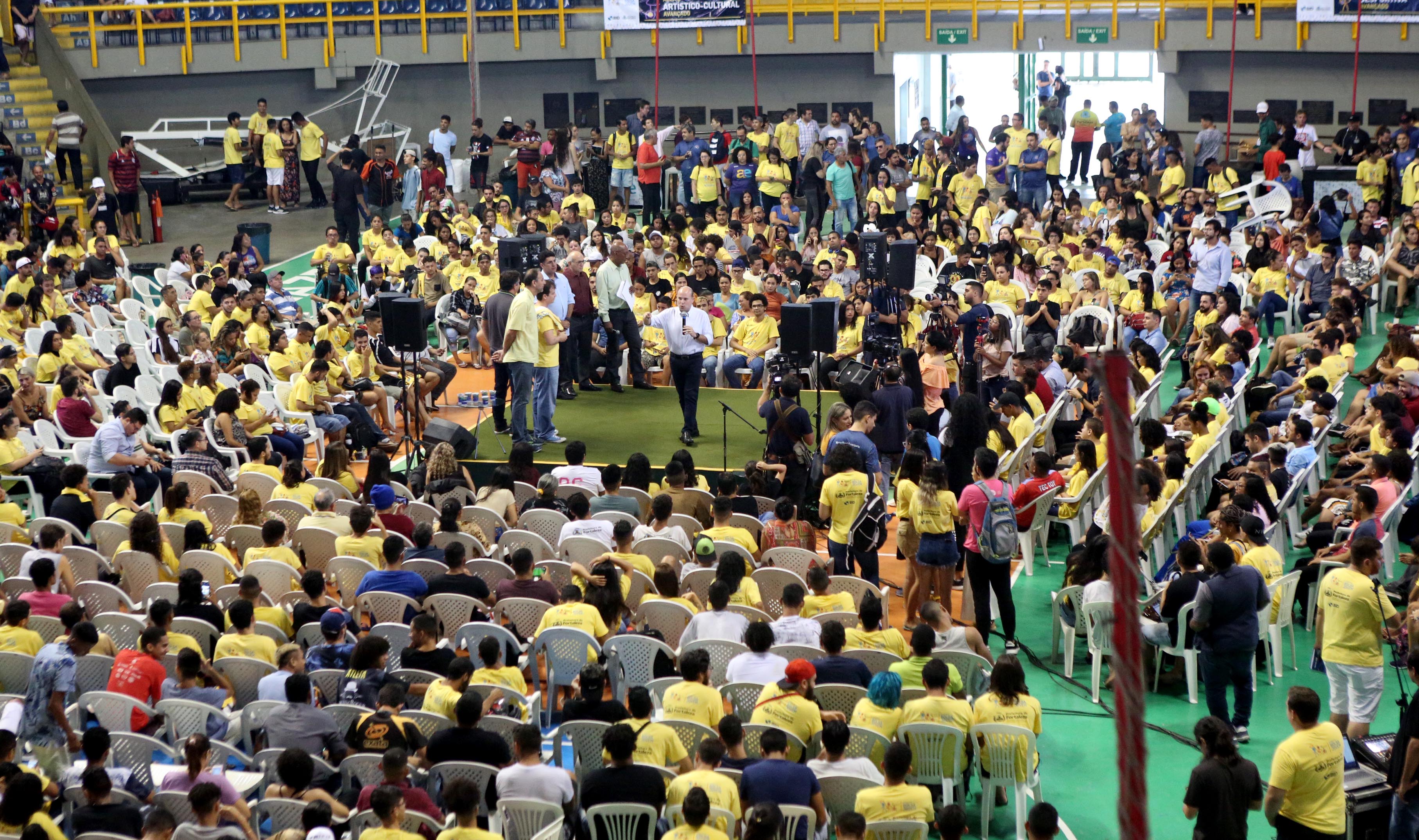 prefeito falando ao microfone em cima de palco com milhates de jovens sentados ao redor assistindo em ginásio